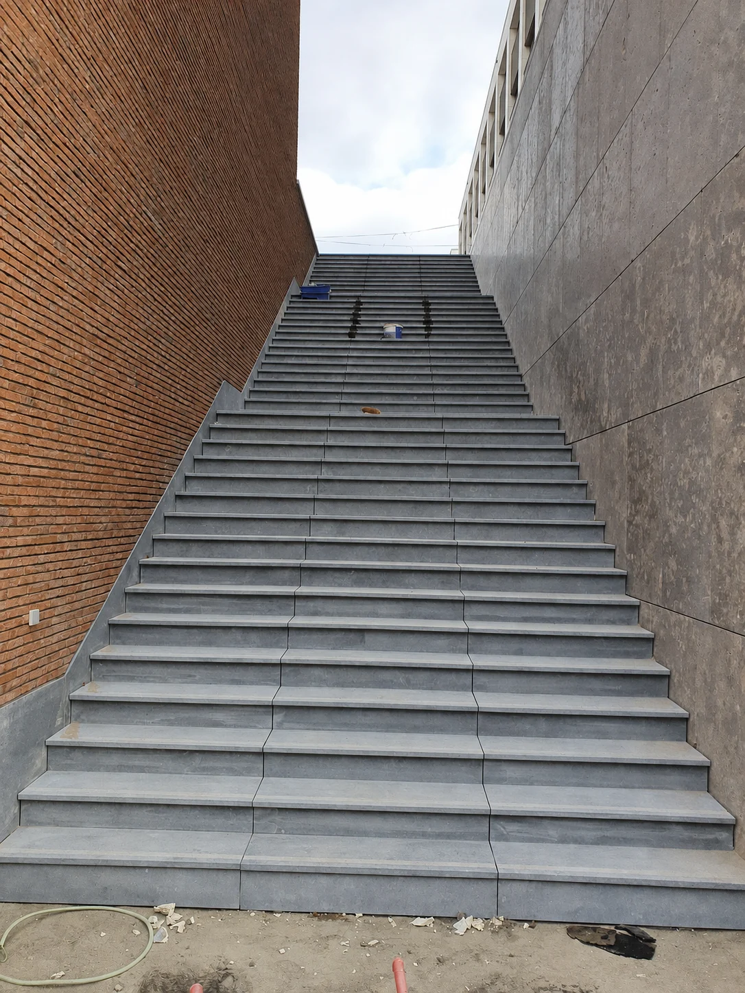 Large escalier extérieur gris entre un mur de briques et un mur revêtu de grands panneaux